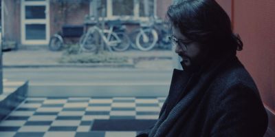 Film still from 'Harmony Was' (2024) by Jon Beaton. A man in a dark coat sits alone in a room with a checkered floor.